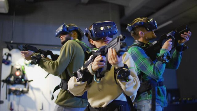 Medium panning shot of group of three young people in virtual reality headsets and other gear playing VR adventure game. Friends with gun controllers standing back to back in defense