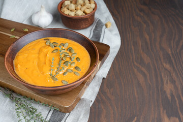 Creamy organic pumpkin soup made of pureed vegetables decorated with seeds and thyme served in bowl on cutting board on dark wooden background with textile, garlic and croutons. Image with copy space