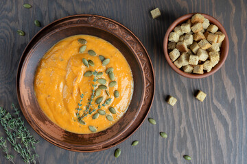 Homemade vegetarian pumpkin soup of orange color made of pureed vegetables cooked on thanksgiving decorated with seeds and thyme served in bowl on dark wooden background with croutons. Horizontal