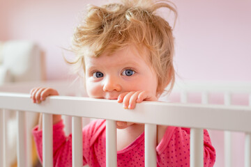 Beautiful blonde one year old baby girl in her cot in pink bedroom looking at camera