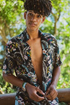 Young African Brazilian man with trendy hairstyle unbuttoning his shirt and looking away in front of an out of focus vegetation background. New normal isolation concept.