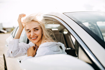 Fototapeta na wymiar Cheerful young blonde woman sitting in car, looking through an open window with wind in hair.