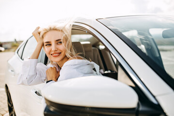 Happy young blonde woman sitting in car, looking through an open window with wind in hair.