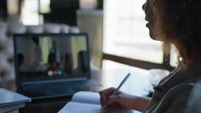 modern teaching, young female student worker taking part in remote video chat conference raises her hand and makes notes in scratchpad while sitting at tables in restaurant by window
