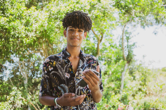 Young African Brazilian man with trendy hairstyle listening to music on his smartphone in front of an out of focus vegetation background. New normal isolation concept.