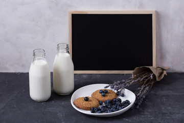 Obraz na płótnie Canvas two glass bottles with milk on a dark background, lavender flowers, homemade cookies and blueberries on a white plate, chalk board for inscriptions. Village breakfast, daily schedule