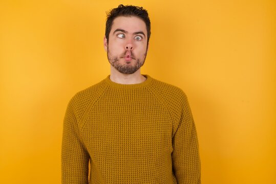 Young man wearing casual sweater and over isolated yellow background making fish face with lips, crazy and comical gesture. Funny expression.