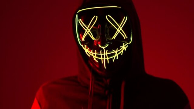 A man in a mask in neon light on a red background. Anonymous shakes his head, halloween mask, unknown horror movie character, masked villain