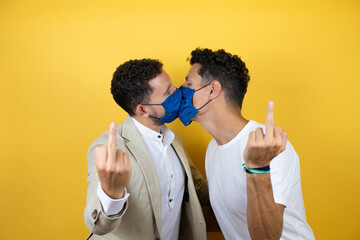 Young gay couple of two men wearing casual clothes and mask over isolated yellow background kissing...