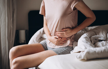 Young woman suffering from strong abdominal pain, casual style indoor shoot. Female sitting on bed...