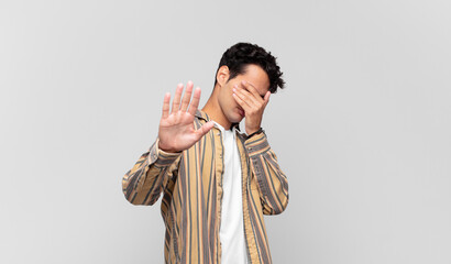 young handsome man covering face with hand and putting other hand up front to stop camera, refusing...