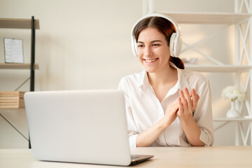 Online communication. Videochat conference. Remote job. Coaching training. Happy woman in white headphones and shirt looking at laptop home workplace interior background.