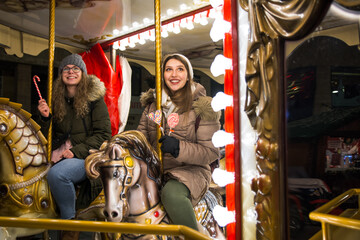 Two female friends riding on the carousel with horses at the winter festival
