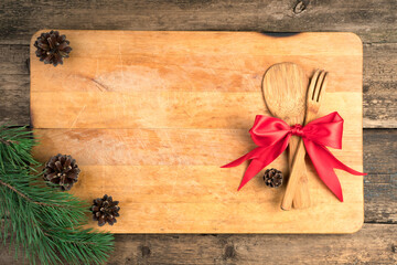 Worn Board with wooden Cutlery tied with a red bow, pine cones and branches on a brown wooden background. The view from the top. The concept of New year and Christmas.