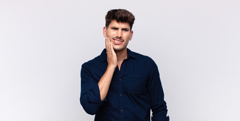 young handsome man holding cheek and suffering painful toothache, feeling ill, miserable and...