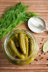 Open glass jar with pickled cucumbers on a wooden table with spices and a fork lying nearby. Concept of cooking or pickling seasonal vegetables.