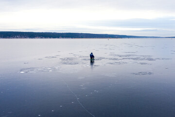 A lone fisherman catches fish from the frozen surface of the river. Next to it is an ice drill. Shooting from a drone.