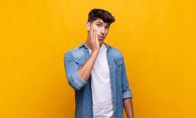 young handsome man feeling shocked and astonished holding face to hand in disbelief with mouth wide...