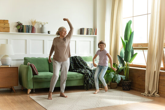Overjoyed barefoot elderly female grandma or babysitter dancing with cute girl on floor carpet at cozy home. Excited grandmother having fun with small child granddaughter in living room.