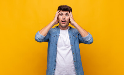 young handsome man feeling horrified and shocked, raising hands to head and panicking at a mistake