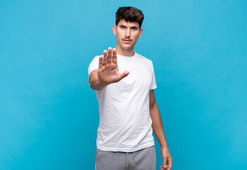 young handsome man looking serious, stern, displeased and angry showing open palm making stop...