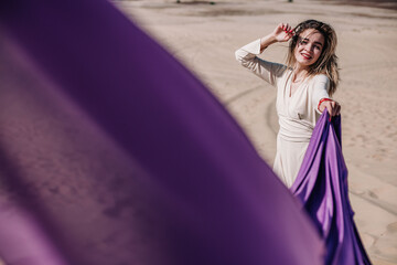 A young, slender girl in a beige dress with purple cloth in her hands posing in the desert in the...