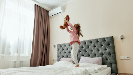 A small girl enjoys spending time holding a teddybear and staying on a huge bed barefoot