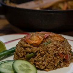 A compact fried rice mound on a white plate with three red shrimps on the top. This is a Thai dish cooked from jasmine rice and vegetables and served with scallions, sliced cucumber and lime.