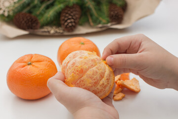 children's hands are cleaning a tangerine, against the background of blurred Christmas tree branches. Close-up photo. Idea - New Years Eve, tangerine flavor, healthy baby food, holiday anticipation