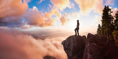 Adventurous Female Hiker on top of a mountain covered in clouds during a vibrant summer sunset. Dramatic Sky Artistic Render. Top of St Mark's Summit, West Vancouver, British Columbia, Canada.