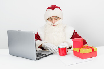 Santa Claus is sitting at his desk, drinking coffee and working at the computer. There are Christmas gifts on the table.