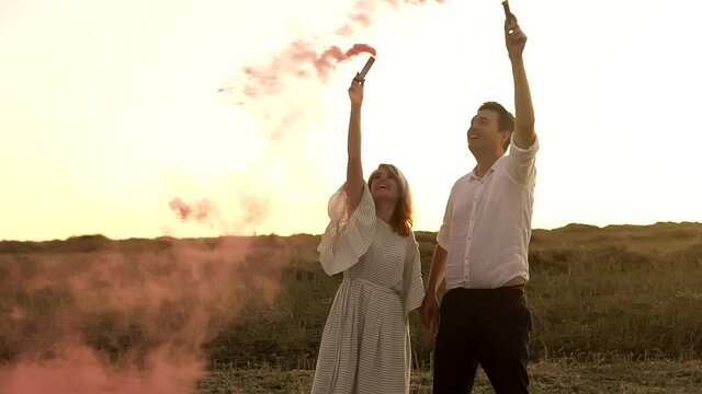 Slowmo Of Man And Woman Who Are Standing In Field And Waving Smoke Bombs On Gender Reveal Party. Young Caucasian Couple Is Expecting Baby Girl. They Are Kissing And Smiling In Summer Meadow On Sunset.