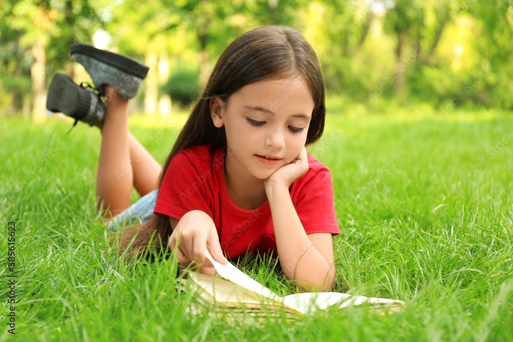 Wall mural Cute little girl reading book on green grass in park