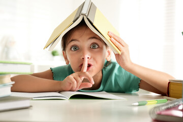 Little girl with book on her head doing homework at table indoors