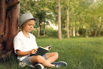 Cute little boy reading book on green grass near tree in park