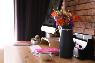 Stylish vase with fresh flowers and cup on wooden table at workplace