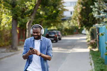 Young Afro-American man in headphones listening music on smart phone using music app. Portrait of smiling guy in earphones and mobile phone outdoors. Walking in the street, relaxation, leisure.