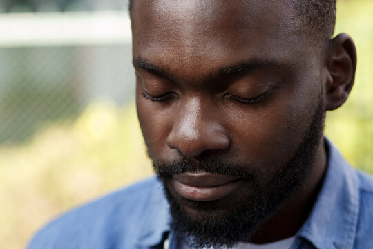 Close up portrait of serious African American man with closed eyes. Handsome young calm man in blue denim shirt meditating on the street.