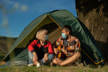 Man and woman in medical masks sing a song to guitar, sitting in a tent. Family is on camping trip, in quarantine. Pandemic, covid19.