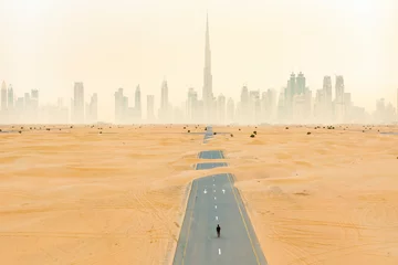 Poster View from above, stunning aerial view of an unidentified person walking on a deserted road covered by sand dunes with the Dubai Skyline in the background. Dubai, United Arab Emirates. © Travel Wild
