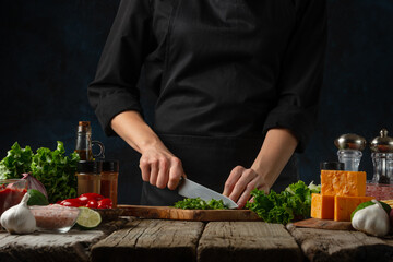 Professional chef in black uniform cuts with knife parsley on chopped board for cooking filling for tacos. Backstage of preparing traditional mexican recipe. Concept of tasty food. Dark background.