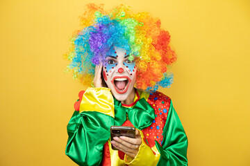 Clown standing over yellow insolated yellow background chatting with her phone and surprised