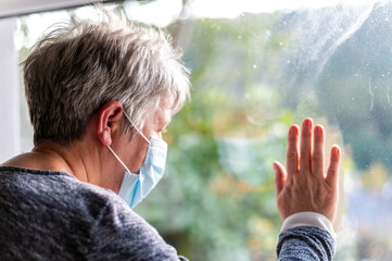 Senior woman wearing protective face mask, looking Out Window, with Covid Virus cells outside of window. Stay Safe stay home concept. shallow focus on face