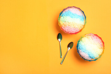 Rainbow shaving ice in glass dessert bowls and spoons on orange background, flat lay. Space for text