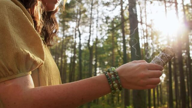 occult science and supernatural concept - young woman or witch with smoking white sage performing magic ritual in forest
