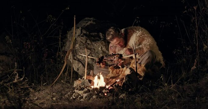 Hungry barbarian eating meat near campfire. Hungry man eating fried meat greedily while resting near bonfire at dark night in nature