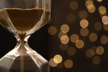 Hourglass with flowing sand against blurred lights, closeup. Christmas countdown