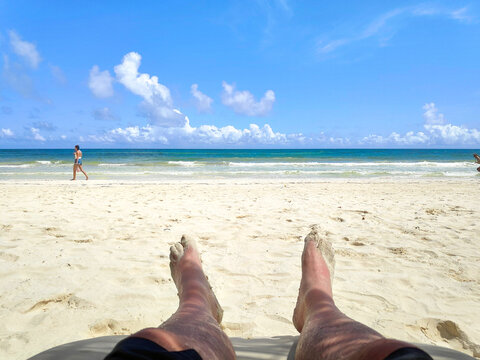 Los pies de un hombre cauc&aacute;sico recostado en un camastro en la playa frente al mar caribe con un cielo azul como fondo.