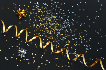 Confetti stars, gold streamer and bows on black background, flat lay. Christmas celebration