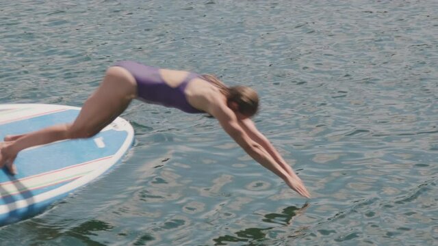 Full shot of athletic woman standing on paddle board and diving underwater with splashes